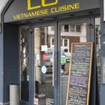 Restaurant LU Vietnamese Eingangsbereich zum Restaurant ist schwarz gestrichen und in gelber Schrift steht der Restaurantname über der Eingangstür. Eine Kreidetafel lehnt an der Hauswand und gibt Einblick auf das Menü.The entrance area to the restaurant is painted black, and the restaurant name is written in yellow letters above the entrance door. A chalkboard leans against the wall of the building, providing a glimpse of the menu.