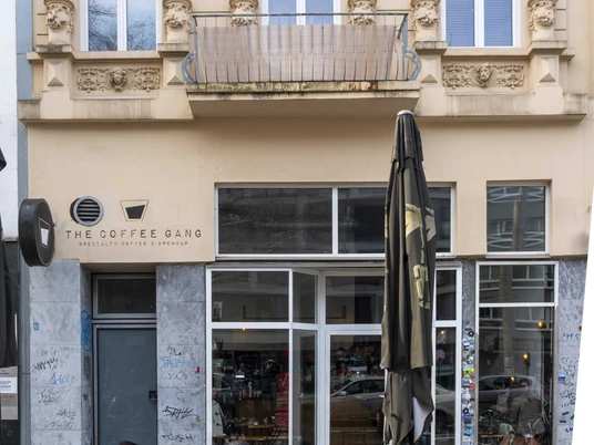The Coffee Gang Die Fassade eines eleganten Altbaus mit ornamentierten Fenstern beherbergt das Café „The Coffee Gang“. Im Vordergrund stehen Holztische und Stühle, einige Gäste unterhalten sich im Freien. The facade of an elegant historic building with ornately decorated windows houses the café "The Coffee Gang". Wooden tables and chairs stand in the foreground, with some guests chatting outside.