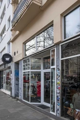 The Coffee Gang Eingang des modernen Cafés "The Coffee Gang" in Köln. Eine breite Glasfront, flankiert von Graffititags auf Naturstein, lässt viel Licht in den Innenraum fallen. Außen stehen Fahrräder geparkt, während das Umfeld von Wohngebäuden mit schlichter Architektur geprägt ist.