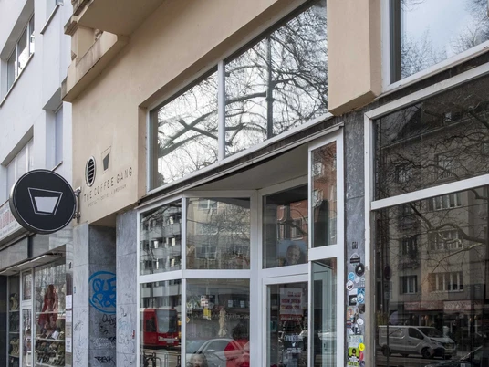 The Coffee Gang Eingang des modernen Cafés "The Coffee Gang" in Köln. Eine breite Glasfront, flankiert von Graffititags auf Naturstein, lässt viel Licht in den Innenraum fallen. Außen stehen Fahrräder geparkt, während das Umfeld von Wohngebäuden mit schlichter Architektur geprägt ist.Entrance to the modern café "The Coffee Gang" in Cologne. A wide glass facade, flanked by graffiti tags on natural stone, allows ample light into the interior space. Bicycles are parked outside, while the surroundings are characterized by residential buildings with simple architecture.