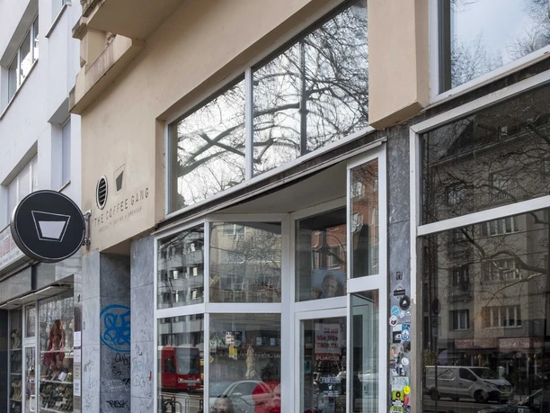 The Coffee Gang Eingang des modernen Cafés "The Coffee Gang" in Köln. Eine breite Glasfront, flankiert von Graffititags auf Naturstein, lässt viel Licht in den Innenraum fallen. Außen stehen Fahrräder geparkt, während das Umfeld von Wohngebäuden mit schlichter Architektur geprägt ist.
