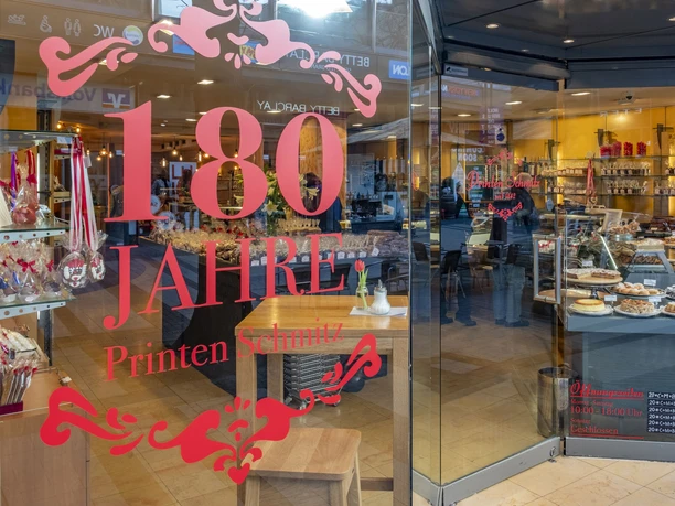 Cafe Printen Schmitz Im Schaufenster des Cafés Printen Schmitz reflektiert die Aufschrift "180 Jahre" die traditionsreiche Jubiläumsfeier, während drinnen vielfältige Backwaren einladend präsentiert werden.