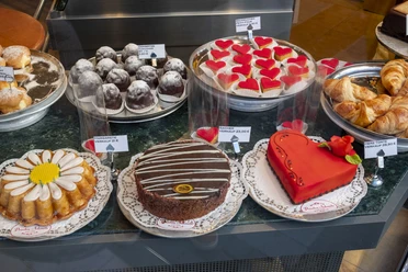Cafe Printen Schmitz An inviting café display case with a selection of artfully decorated confections. On display are a variety of cakes and pies, including a striking heart-shaped pie with red decoration, presented on a white doily.