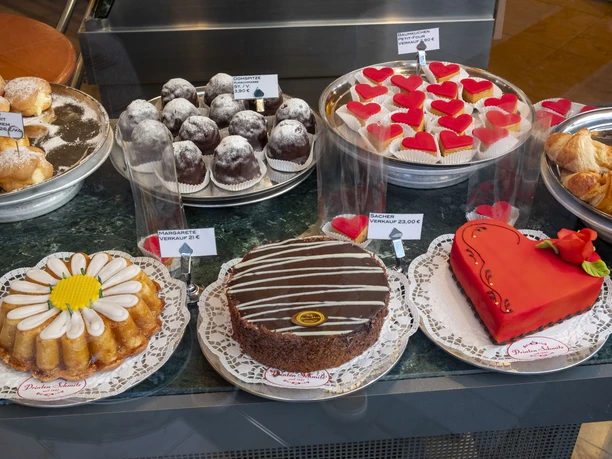 Cafe Printen Schmitz Eine einladende Vitrine eines Cafés mit einer Auswahl an kunstvoll dekorierten Süßwaren. Zu sehen sind eine Vielzahl von Kuchen und Torten, darunter eine auffällige herzförmige Torte mit einer roten Verzierung, die auf einer weißen Tortenspitze präsentiert wird.