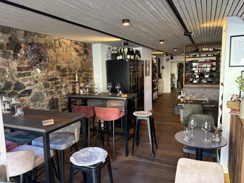 Herford-Weinbar 1575-Gastrobereich-Hartendorf (6).jpeg Ein stilvoller Innenbereich mit Holztischen und farbigen Barhockern vor einer rustikalen Steinwand.