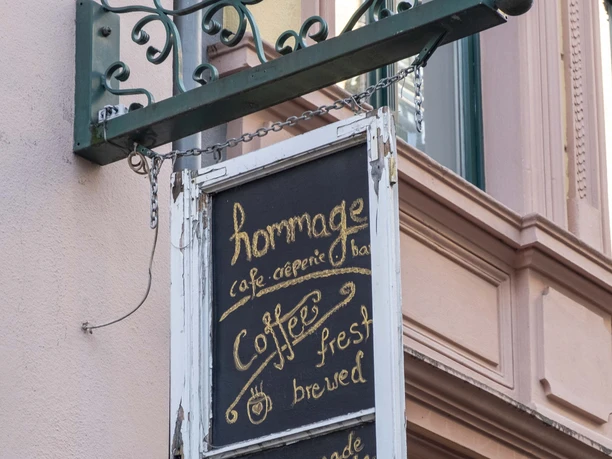 Cafe Hommage Eine Kreidetafel im Shabby Chic Stil ist an einem Metallgestell an der Hausfassade befestigt.