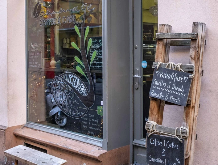 Cafe Hommage Einladendes Fenster mit kleiner Holzbank davor, daneben eine Kreidetafel an einer Holzleiter befestigt.Inviting window with a small wooden bench in front of it, next to it a chalk board attached to a wooden ladder.