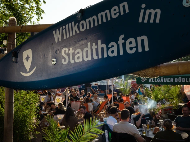 Stadtstrand des Stadthafen Leipzig - Wasserstadt Leipzig Blaues Willkommensschild vom Stadthafen Leipzig in Form eines Surfbrettes am Stadtstrand, darunter viele Menschen in Liegestühlen sitzend, Wasserstadt Leipzig