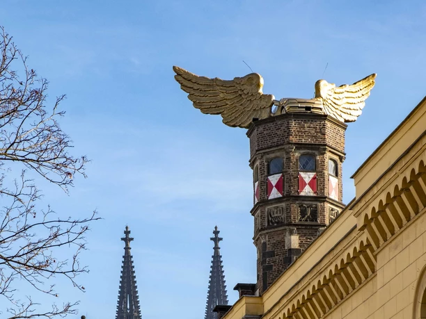 Goldenes Flügelauto Historische Fassade eines Kölner Gebäudes, gekrönt von goldenen Adlerflügeln. Dahinter ragen die Türme des Kölner Doms majestätisch in den blauen Himmel.