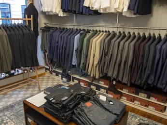 Herrenbude Modegeschäft mit dezenter Beleuchtung, zahlreiche Herren-Anzüge und gefaltete Jeans auf Holztisch.Fashion store with discreet lighting, numerous men's suits and folded jeans on a wooden table.