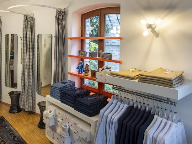 Herrenbude Ein stilvoll eingerichteter Modeladen mit hölzernem Fußboden, in dem ordentlich gefaltete Kleidung und stilvoll platzierte Dekorationen zu sehen sind. Das Bild zeigt einen eleganten Innenraum mit sorgfältig arrangierten Textilien und geschmackvoller Beleuchtung.