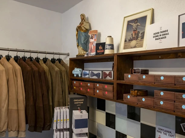 Herrenbude Zu sehen sind Jacken an einem Kleiderständer, Accessoires wie Fliegen und Tücher in Regalen sowie Retro-Dekorationen und moderne Designartikel.