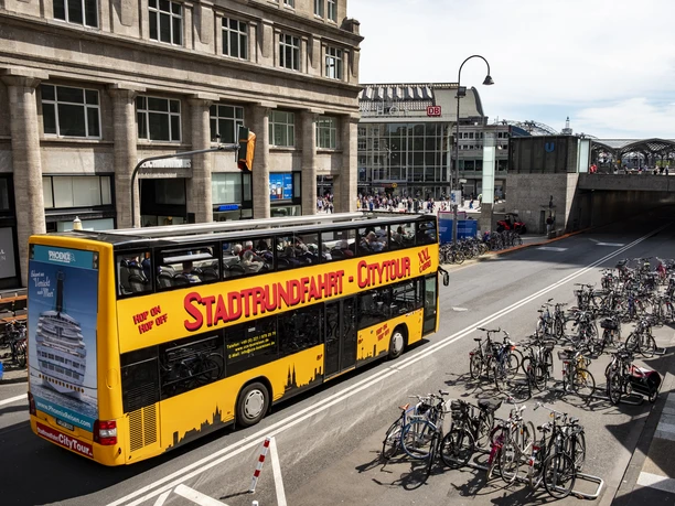 Kölner CityTour Ein markanter, gelber Doppeldeckerbus für Stadtrundfahrten fährt an der Kölner Altstadt vorbei. Zahlreiche Fahrräder stehen in einem eigens dafür vorgesehenen Bereich.