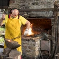 Aufschlag am Oelchenshammer Ein Schmied bearbeitet heißes Metall auf einem Amboss in einer Werkstatt mit offenem Kaminfeuer.