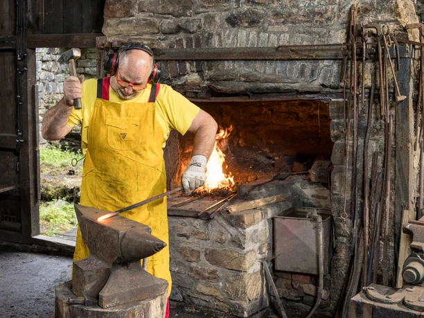 Aufschlag am Oelchenshammer Ein Schmied bearbeitet heißes Metall auf einem Amboss in einer Werkstatt mit offenem Kaminfeuer.
