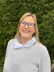 Stadtführerin Gudrun Berger Porträt einer lächelnden Frau mit Brille, hellem Pullover und Schal, vor einem Hintergrund aus grünem Laub.