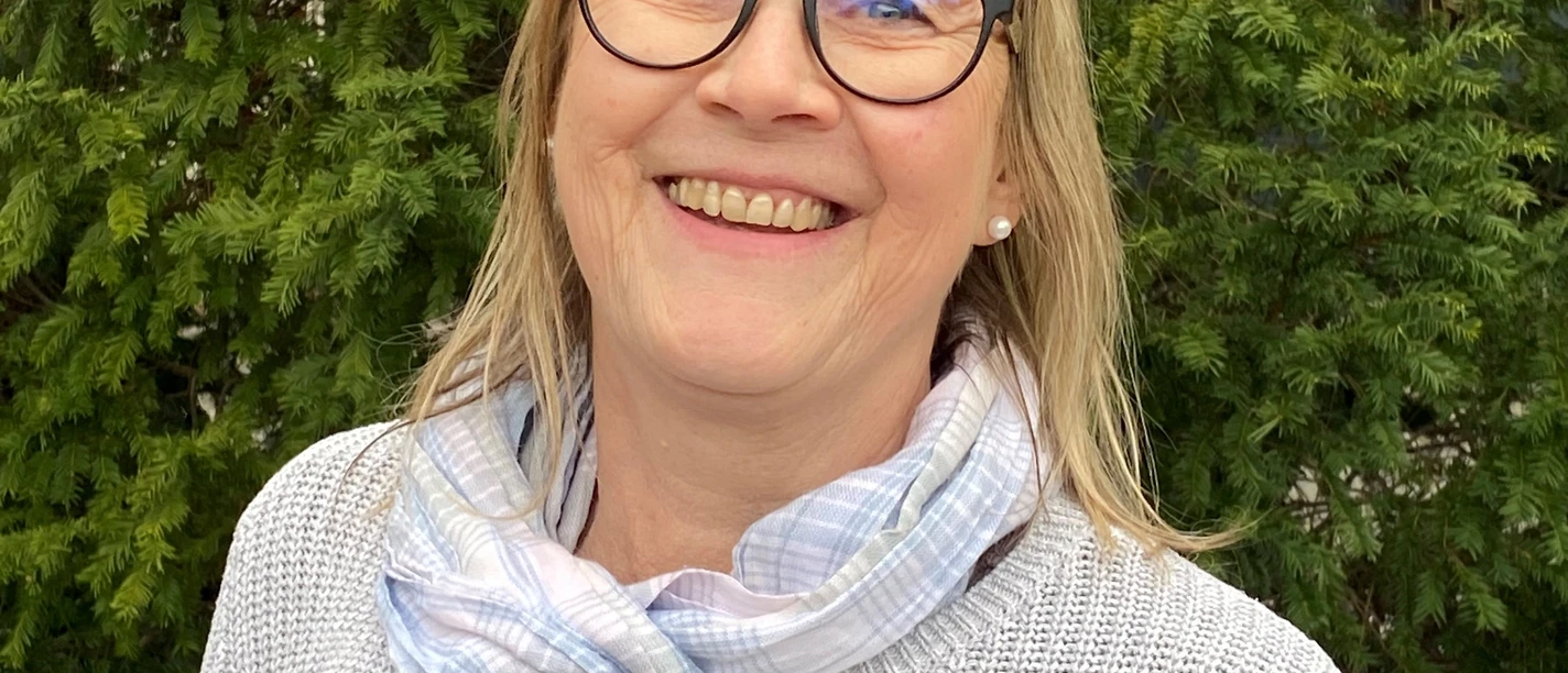 Stadtführerin Gudrun Berger Porträt einer lächelnden Frau mit Brille, hellem Pullover und Schal, vor einem Hintergrund aus grünem Laub.