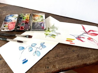 Aquarellmalen Aquarellfarben und Pinsel auf einem Holztisch, daneben zwei bemalte Papierblätter.