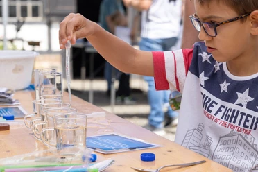 Platsch am Oelchenshammer Ein Junge mit Brille misst in Gläsern mit einer Pipette Flüssigkeit, draußen bei einer Veranstaltung.