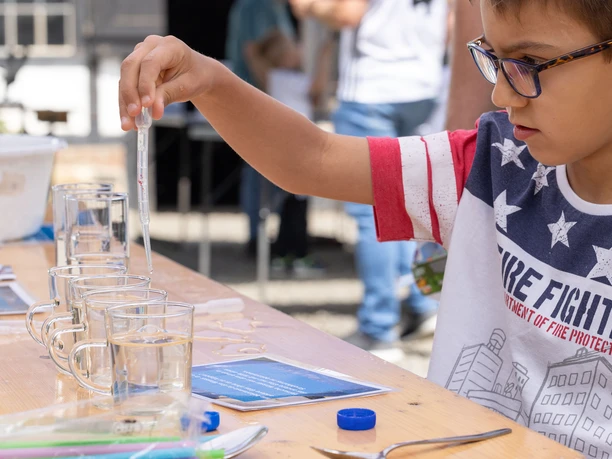 Platsch am Oelchenshammer Ein Junge mit Brille misst in Gläsern mit einer Pipette Flüssigkeit, draußen bei einer Veranstaltung.