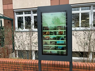 A digital information kiosk against a brick wall in front of a building, surrounded by shrubs.