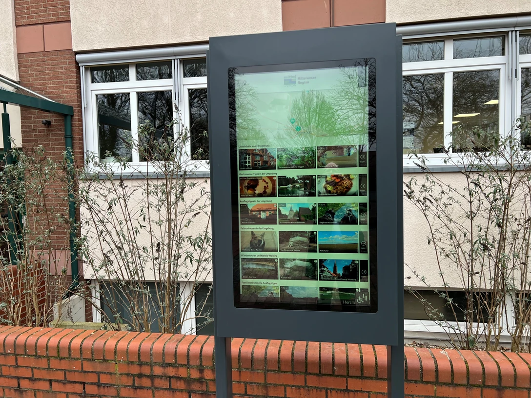 Infoterminal Steimbke Digitales Infoterminal an einer Ziegelmauer vor einem Gebäude, von Sträuchern umgeben.