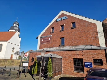 AußenansichtKöstersCC-BY-SA_Teutoburger_Wald_Stadt_Schloss_Holte-Stukenbrock_HotelKösters.jpg Hotel Kösters in Backsteinoptik, rechts ein Parkplatzschild, links die Kirche mit Zwiebelturm.