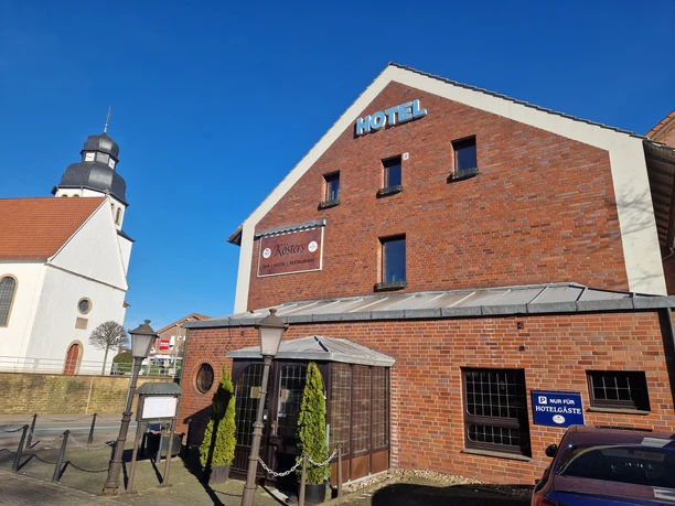 AußenansichtKöstersCC-BY-SA_Teutoburger_Wald_Stadt_Schloss_Holte-Stukenbrock_HotelKösters.jpg Hotel Kösters in Backsteinoptik, rechts ein Parkplatzschild, links die Kirche mit Zwiebelturm.