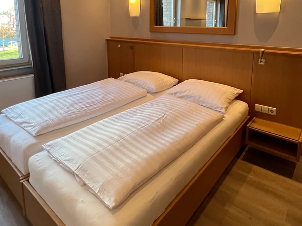 DoppelzimmerCC-BY-SA_Teutoburger_Wald_Stadt_Schloss_Holte-Stukenbrock_HotelKösters.jpeg Helles Doppelzimmer mit einem großen Bett, Holzmöbeln und einem Fenster mit Blick nach draußen.