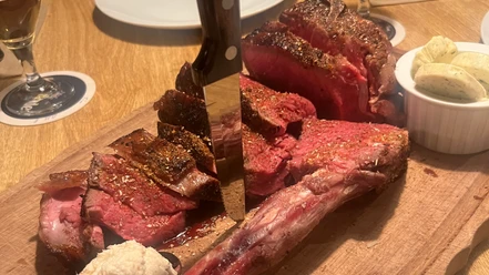 Tomahawk-SteakCC-BY-SA_Teutoburger_Wald_Stadt_Schloss_Holte-Stukenbrock_HotelKösters.jpeg Ein aufgeschnittenes Tomahawk-Steak auf einem Holzbrett, begleitet von Kräuterbutter und Dip.