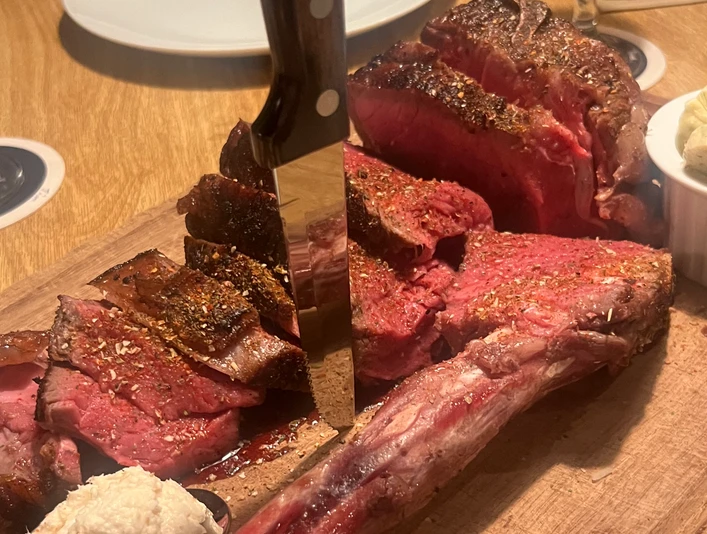 Tomahawk-SteakCC-BY-SA_Teutoburger_Wald_Stadt_Schloss_Holte-Stukenbrock_HotelKösters.jpeg Ein aufgeschnittenes Tomahawk-Steak auf einem Holzbrett, begleitet von Kräuterbutter und Dip.