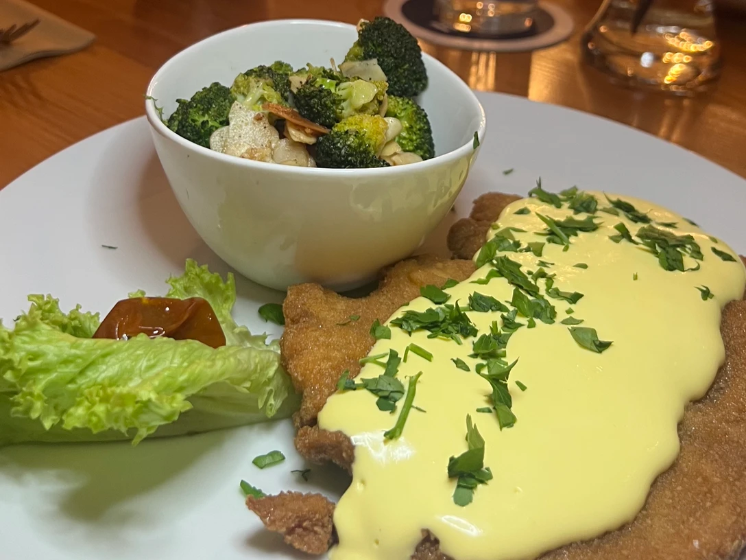 Ein Teller mit Schnitzel, bedeckt mit heller Sauce, Brokkoli-Gemüse und einem Salatblatt.
