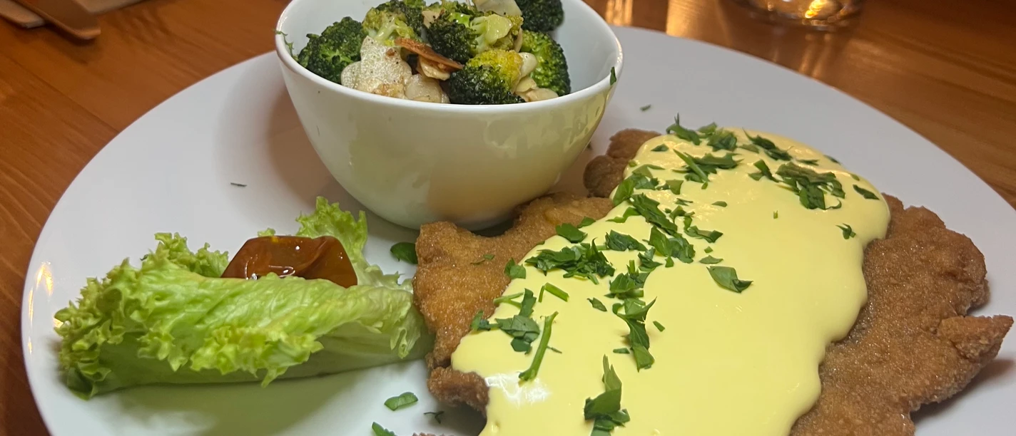 Ein Teller mit Schnitzel, bedeckt mit heller Sauce, Brokkoli-Gemüse und einem Salatblatt.