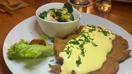 SchnitzelBrokkoliHollandaiseCC-BY-SA_Teutoburger_Wald_Stadt_Schloss_Holte-Stukenbrock_HotelKösters.jpeg Ein Teller mit Schnitzel, bedeckt mit heller Sauce, Brokkoli-Gemüse und einem Salatblatt.