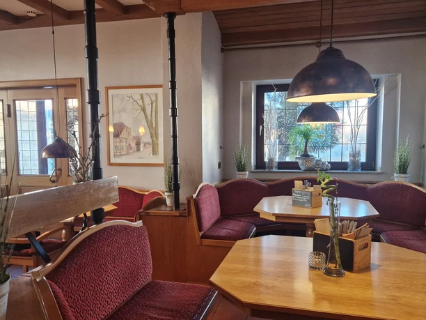 Restaurant1CC-BY-SA_Teutoburger_Wald_Stadt_Schloss_Holte-Stukenbrock_HotelKösters.jpg Restaurantecke mit Holztischen, roten Sitzbänken und Lampen, sowie Fenstern mit Ausblick.