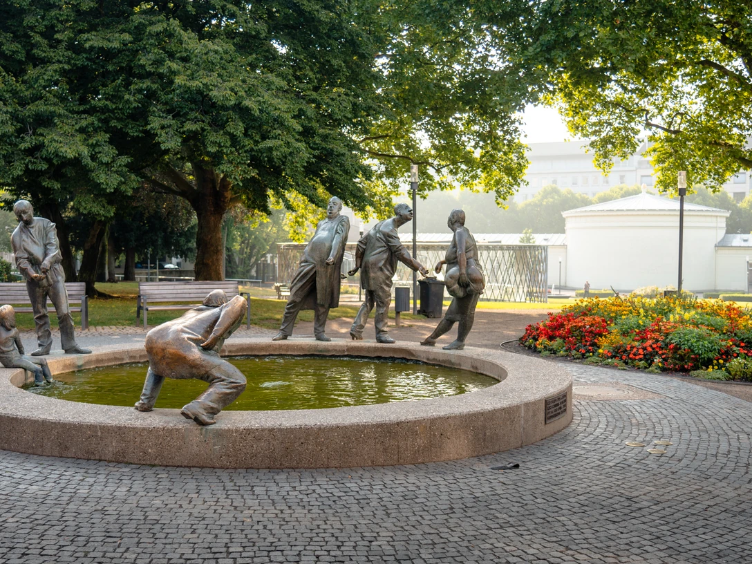 2022-10 Kreislauf des Geldes 2 (c) aachenviews.jpg Die Skulptur „Kreislauf des Geldes“ zeigt sechs bronzene Figuren, die Geiz, Gier, Gönnerhaftigkeit und Bettelei symbolisieren. Er befindet sich vor einem Park.