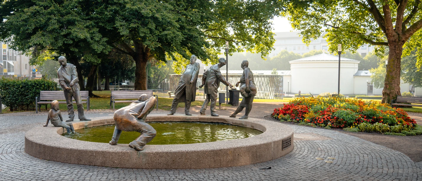 2022-10 Kreislauf des Geldes 2 (c) aachenviews.jpg Die Skulptur „Kreislauf des Geldes“ zeigt sechs bronzene Figuren, die Geiz, Gier, Gönnerhaftigkeit und Bettelei symbolisieren. Er befindet sich vor einem Park.
