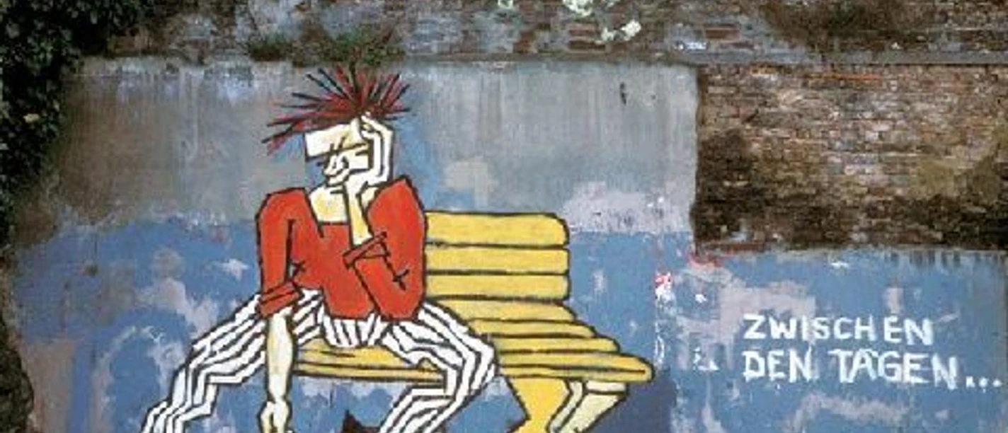 Street Art Führung-Aachener Innenstadt Zwischen den Tagen (c) Regina Weinkauf.jpg Graffiti einer Punk-Person auf einer Bank mit der Aufschrift „Zwischen den Tagen“ vor einer alten Mauer.