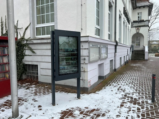 An information kiosk stands in front of a snow-covered sidewalk next to a historic building with white walls.