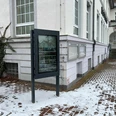 Infoterminal Uchte Ein Infoterminal steht vor einem schneebedeckten Gehweg neben einem historischen Gebäude mit weißen Wänden.