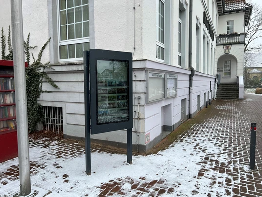 Infoterminal Uchte Ein Infoterminal steht vor einem schneebedeckten Gehweg neben einem historischen Gebäude mit weißen Wänden.