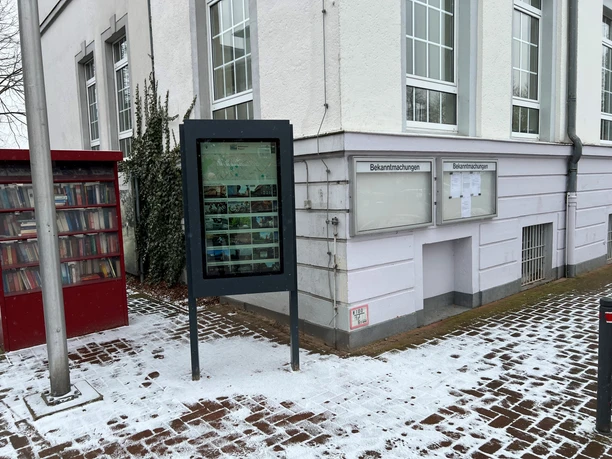 A freestanding information kiosk in front of a white wall with a display case, surrounded by snow and a bookshelf.