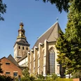 Saint Ursula Sankt Ursula Kirche mit markantem Glockenturm, umgeben von Bäumen, unter klarem Himmel.St. Ursula's Church with its striking bell tower, surrounded by trees, under a clear sky.