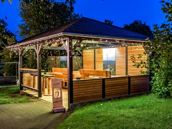 Pavillon vom Landhotel Alte Linde Würgassen Pavillon, umgeben von Bäumen und Rasenfläche, Bestuhlung mit Barhockern und Sitzbänken aus Holz, Lichterketten