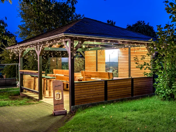 Pavillon vom Landhotel Alte Linde Würgassen Pavillon, umgeben von Bäumen und Rasenfläche, Bestuhlung mit Barhockern und Sitzbänken aus Holz, Lichterketten