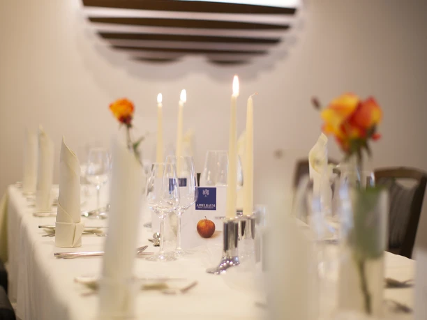 Gedeckter Tisch im Gütsel Ein festlich gedeckter Tisch mit weißen Tischtüchern, Kerzen und orangen Rosen in einem eleganten Raum.