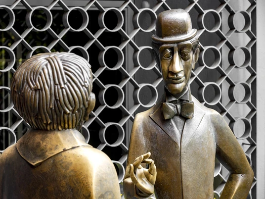 Tünnes and Schäl Monument Das Bild zeigt die berühmten Bronze-Figuren "Tünnes und Schäl" am Willy-Millowitsch-Platz in Köln. Beide Figuren stehen einander gegenüber; Tünnes trägt eine Mütze und Schäl einen Zylinder. Im Hintergrund ist ein kunstvoll geschmiedetes Gitter zu sehen.The picture shows the famous bronze figures "Tünnes and Schäl" at Willy-Millowitsch-Platz in Cologne. Both figures are standing opposite each other; Tünnes is wearing a cap and Schäl a top hat. An artistically forged grille can be seen in the background.