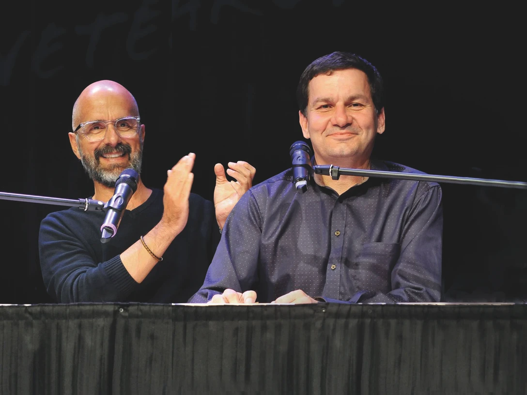 Zwei Männer sitzen fröhlich an einem Tisch auf einer Bühne, sprechen in Mikrofone und lachen.