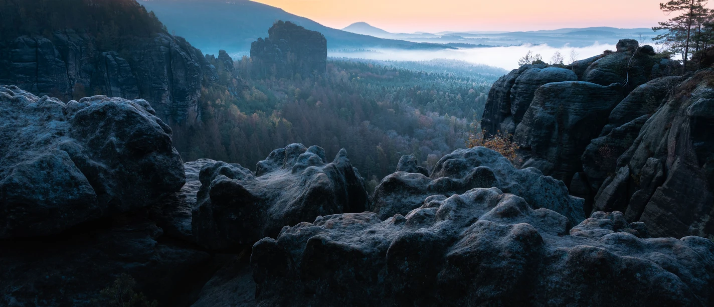 Breite Kluft Felsige Landschaft bei Sonnenaufgang mit Blick auf bewaldete Hügel und Nebel im Tal, im Hintergrund ein klarer Himmel mit orangefarbenem Horizont.