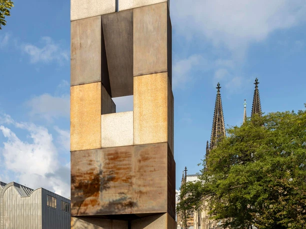 MAALOT Ein beeindruckendes Kunstobjekt aus rostigem Stahl erhebt sich vor der Kulisse des Kölner Doms. Der Turm wirkt monumental und kontrastiert mit dem blauen Himmel und den gotischen Kirchenspitzen.